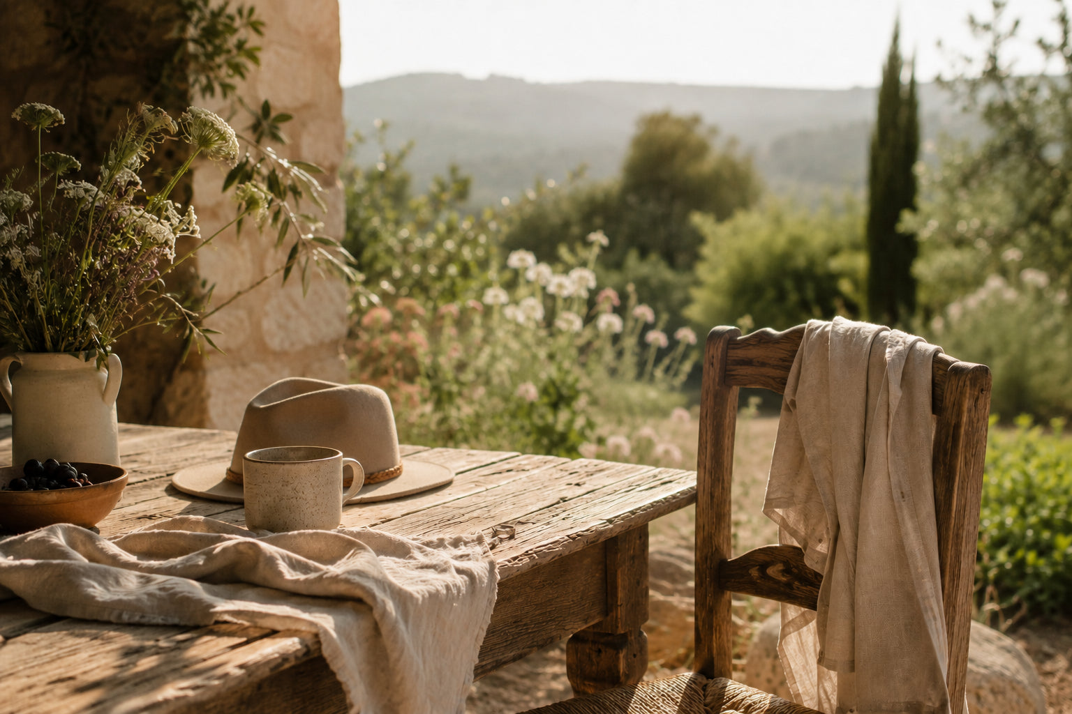 Rustikaler Holztisch mit Leinentuch, Keramiktasse, Wildblumen und Hut auf einer sonnigen Terrasse – inspiriert von der natürlichen Markenwelt von Spitzstock.